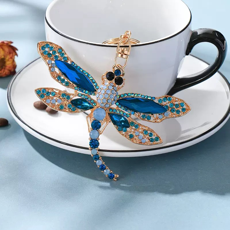Decorative dragonfly keychain with blue gemstones on a saucer next to a cup
